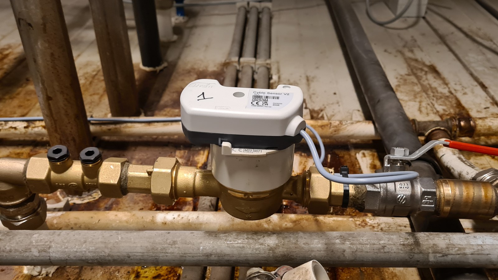 Compteur d'eau équipé d'un capteur LoRaWAN à impulsions fixé sur son corps, entre les vannes d'arrêt et un raccord union, au milieu de tuyauteries d'atelier